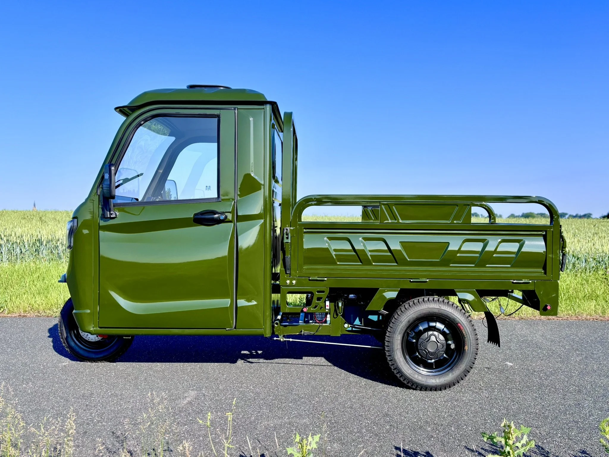 En grøn elektrisk luxcap el-kabinescooter med lad i en mark eller vej mod en blå himmel.