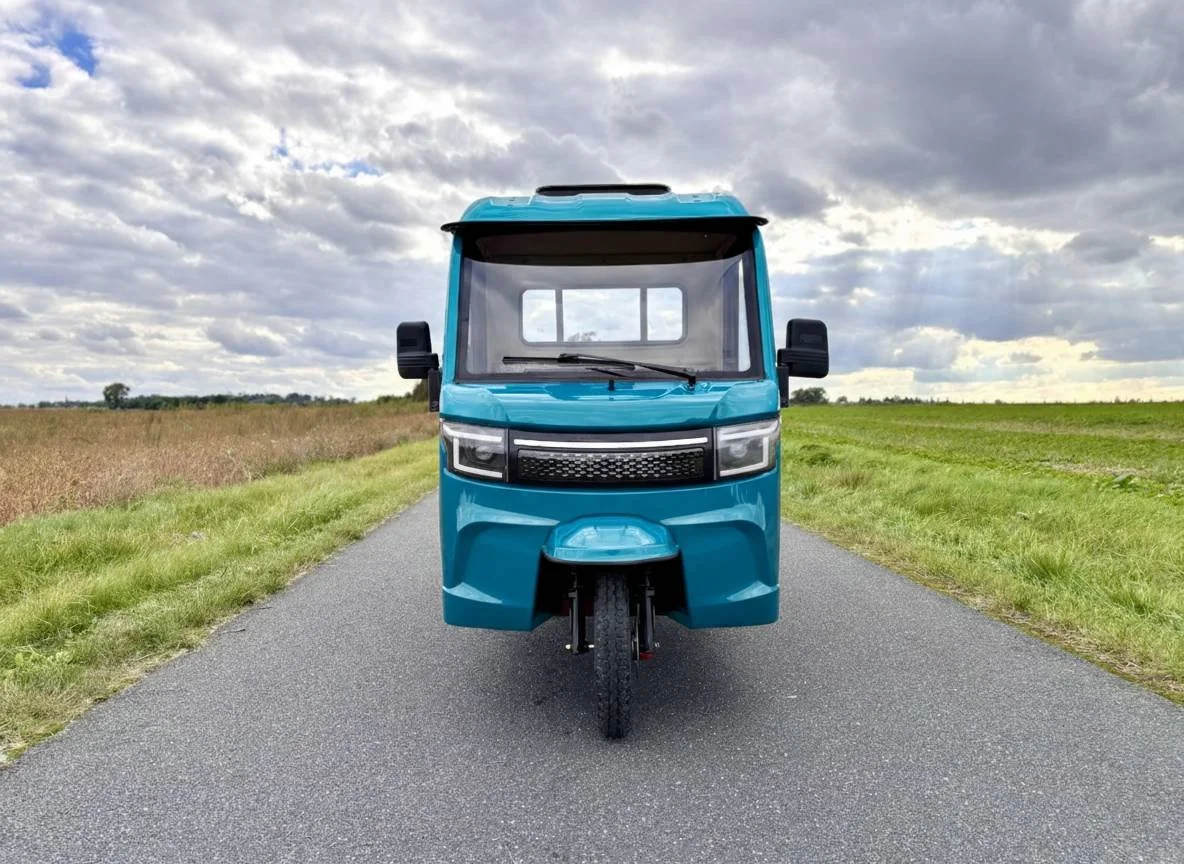 En blå luxcap el-kabinescooter køres mod kameraet på en asfaltvej, omgivet af marker med græs og afgrøder under en overskyet himmel.