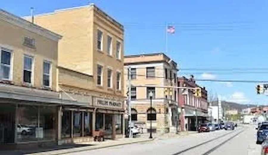 Street scene with historic buildings, cars parked along the curb, and an American flag flying in the background on a clear day.
