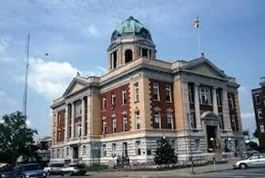 Historic courthouse building with a dome, brick and stone facade, American flag, and parked cars in front
