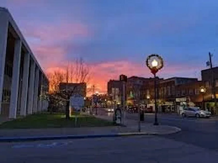 Sunset sky over a small downtown street with buildings, cars, a lamp post with a wreath decoration, and a person walking.