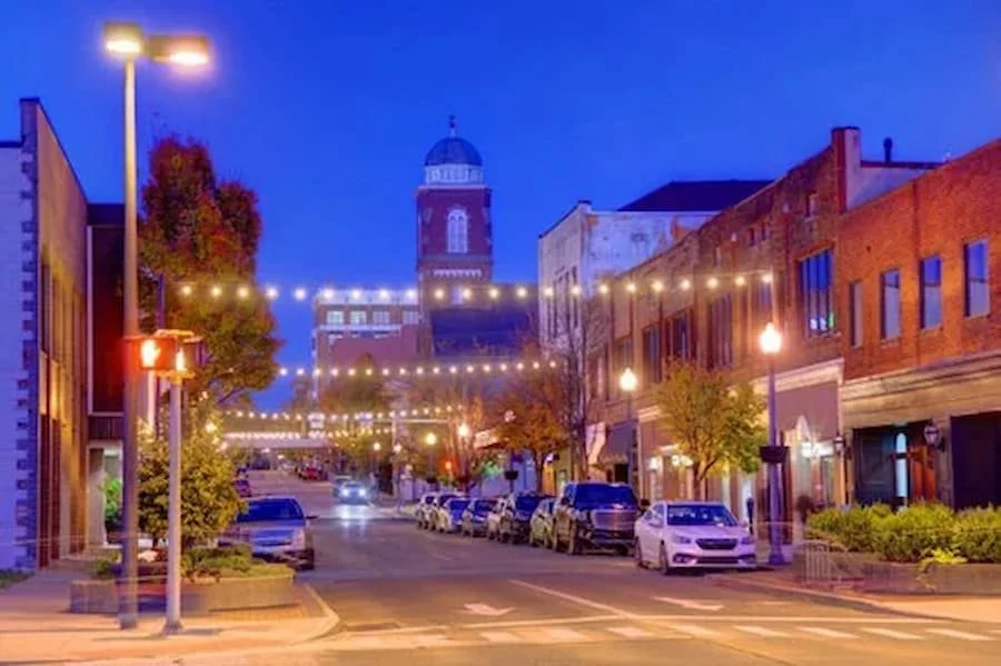 Downtown street at dusk with string lights overhead, parked cars along the street, and historic brick buildings. A clock tower is visible in the background.
