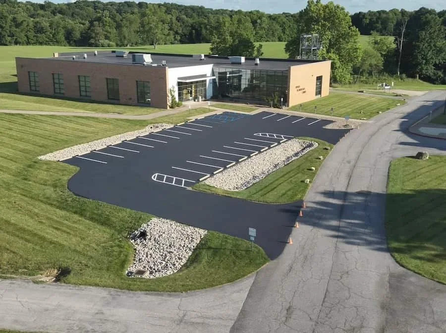 A modern building with a large parking lot, surrounded by green grass and trees, with a clear sky in the background.