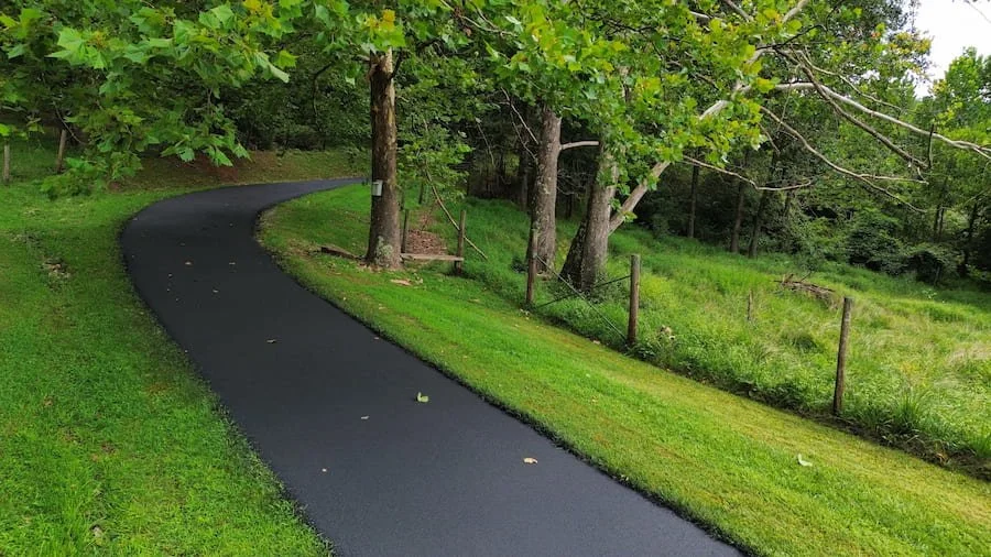 A winding asphalt pathway through a grassy area with trees and a fence.