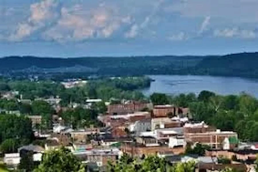 A small town with brick buildings and green trees, next to a river and rolling hills or mountains in the background under a partly cloudy sky.