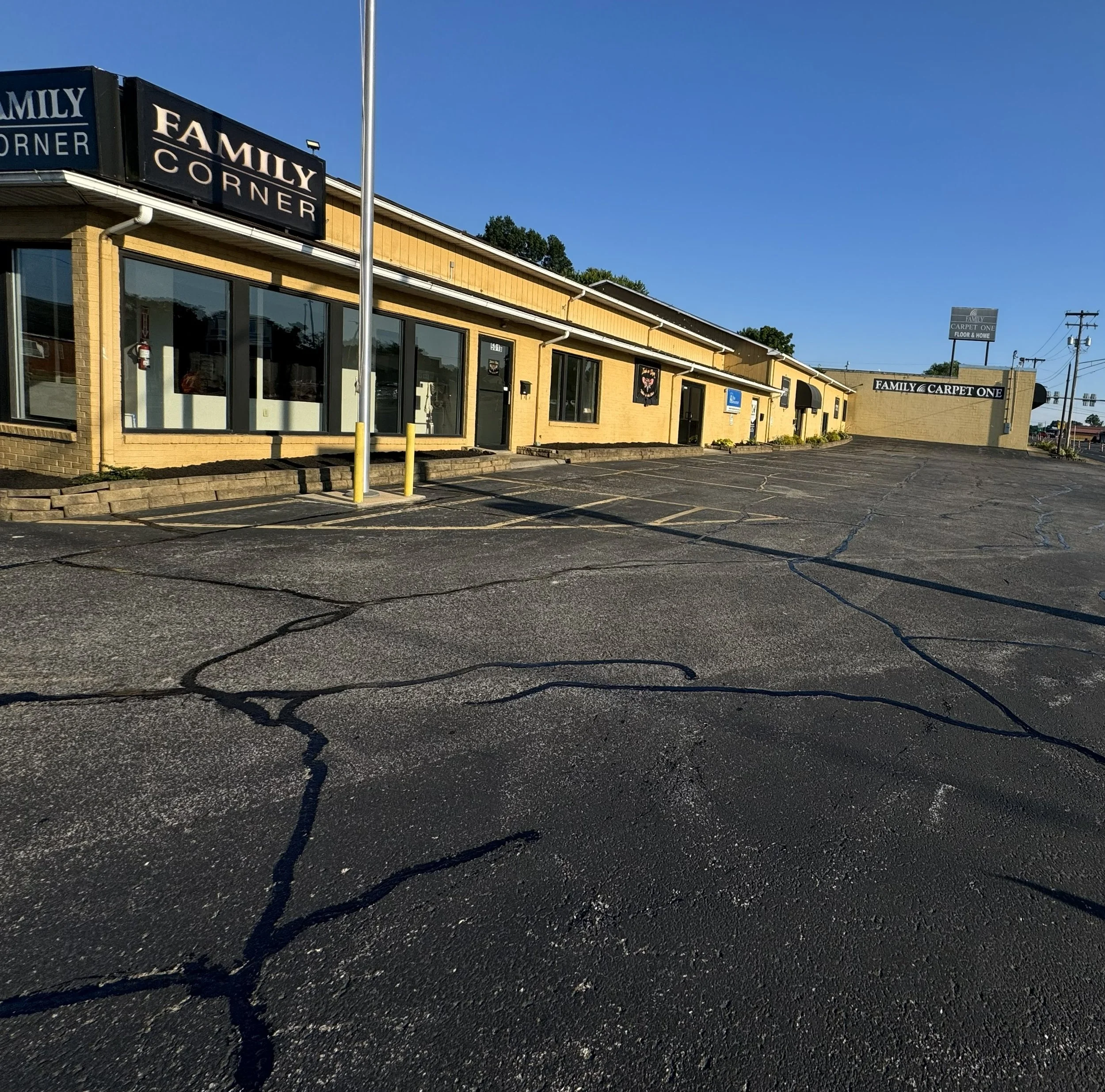 Empty parking lot in front of a beige commercial building with signs reading "Family Corner" and "Family Carpet One," under a clear blue sky.