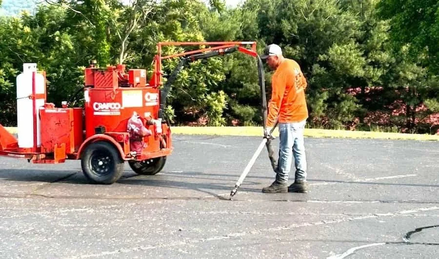A worker using a line striping machine on the asphalt of a parking lot. The machine is red, and the worker is wearing an orange shirt and white cap.