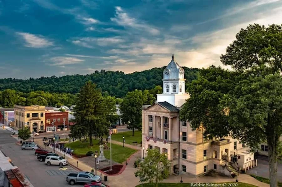 A small town square with trees, parked cars, a historic building with a clock tower, and a hilly, forested backdrop during sunset.