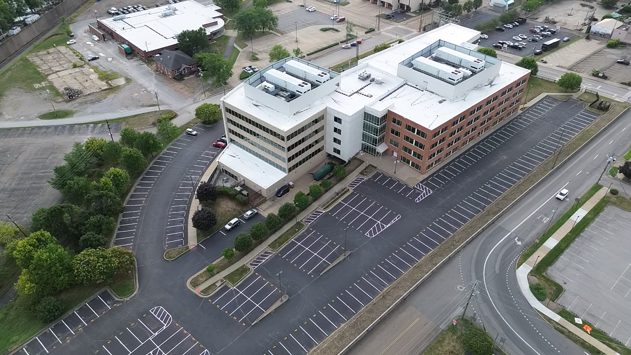 An aerial view of a multi-story office building with a parking lot and surrounding streets, with some trees and adjacent empty lots.