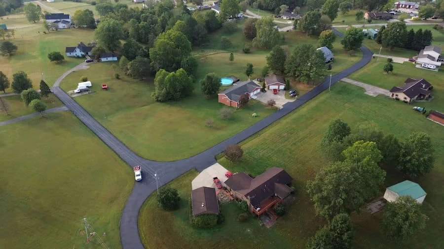 Aerial view of a suburban neighborhood with winding asphalt roads, green lawns, trees, houses, a swimming pool, and parked vehicles.