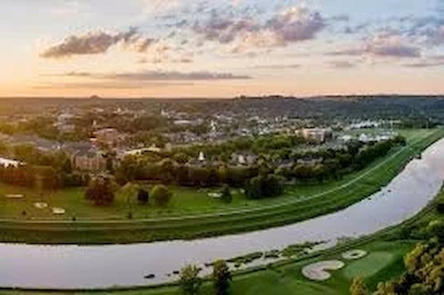 A scenic view of a city with a river in the foreground, a park with trees and open grassy areas, and a sunset sky with clouds.