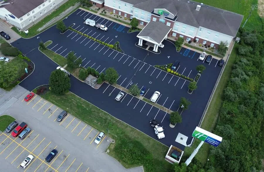An aerial view of a hospital parking lot with several parked cars, surrounding trees, and a building with a covered entrance. The parking lot has dedicated spaces for handicapped parking and is adjacent to another lot with mostly empty spaces.