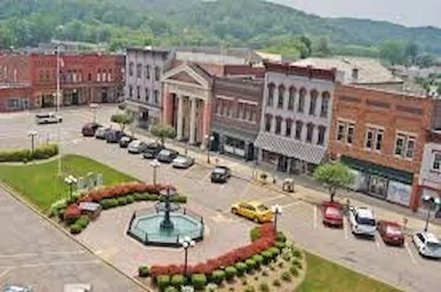 A small town square with a fountain and flower beds, surrounded by brick buildings and parked cars, with green hills in the background.