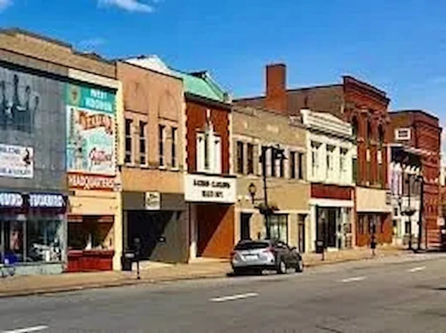 A row of small businesses and storefronts on a street with parked cars, under a clear blue sky.