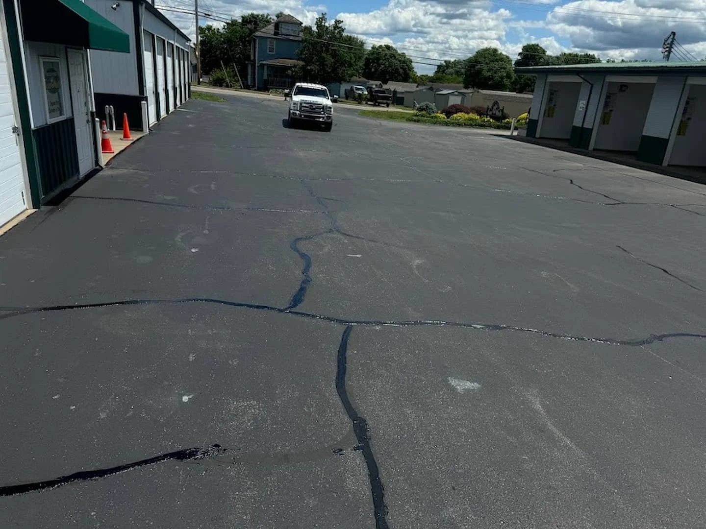 A parking lot with cracks in the asphalt, a white pickup truck parked at the distance, and buildings on either side with a blue house in the background and partly cloudy sky.
