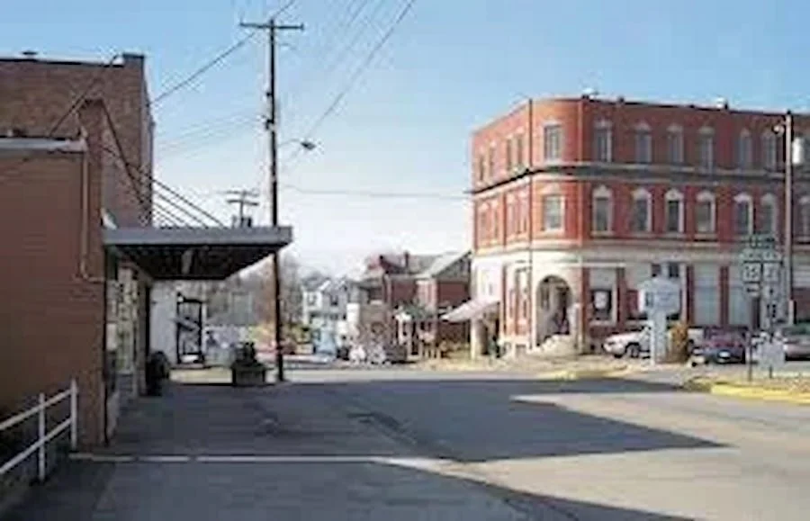 A small town intersection with brick buildings, parked cars, utility poles, and a clear blue sky.