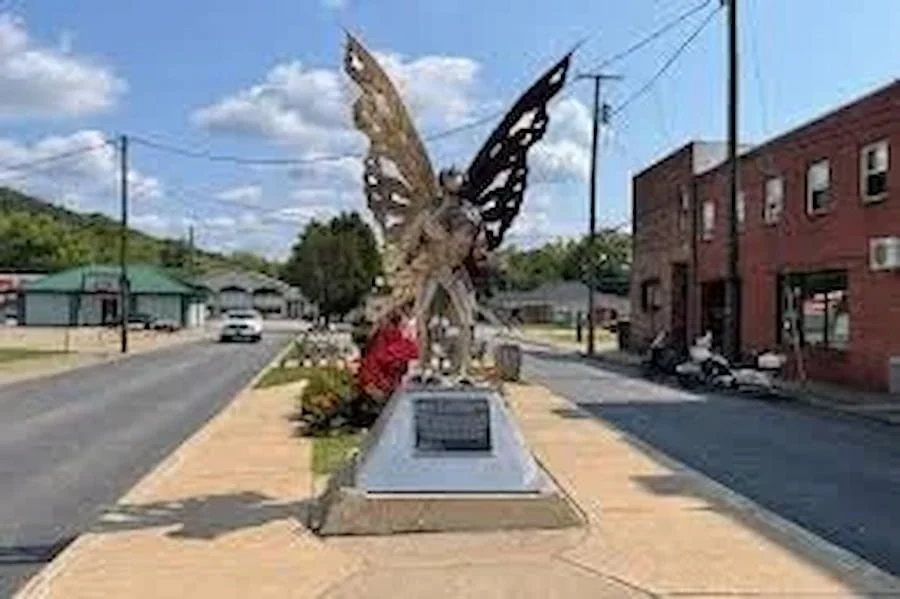A large butterfly sculpture on a display platform in a small town street, surrounded by buildings and parked motorcycles.