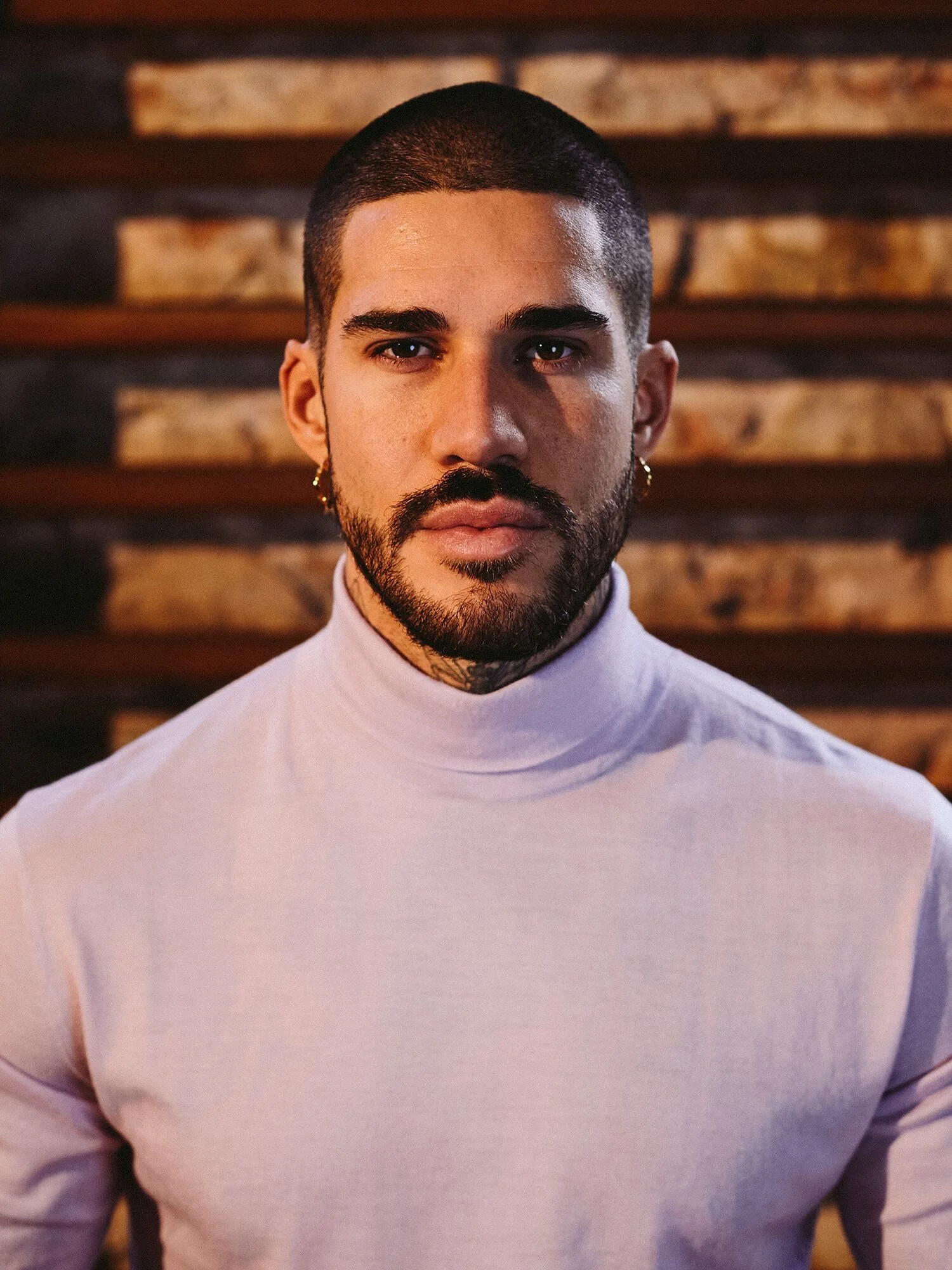 A man with short dark hair, a beard, and earrings, wearing a high-neck light-colored shirt, standing in front of a wooden wall.