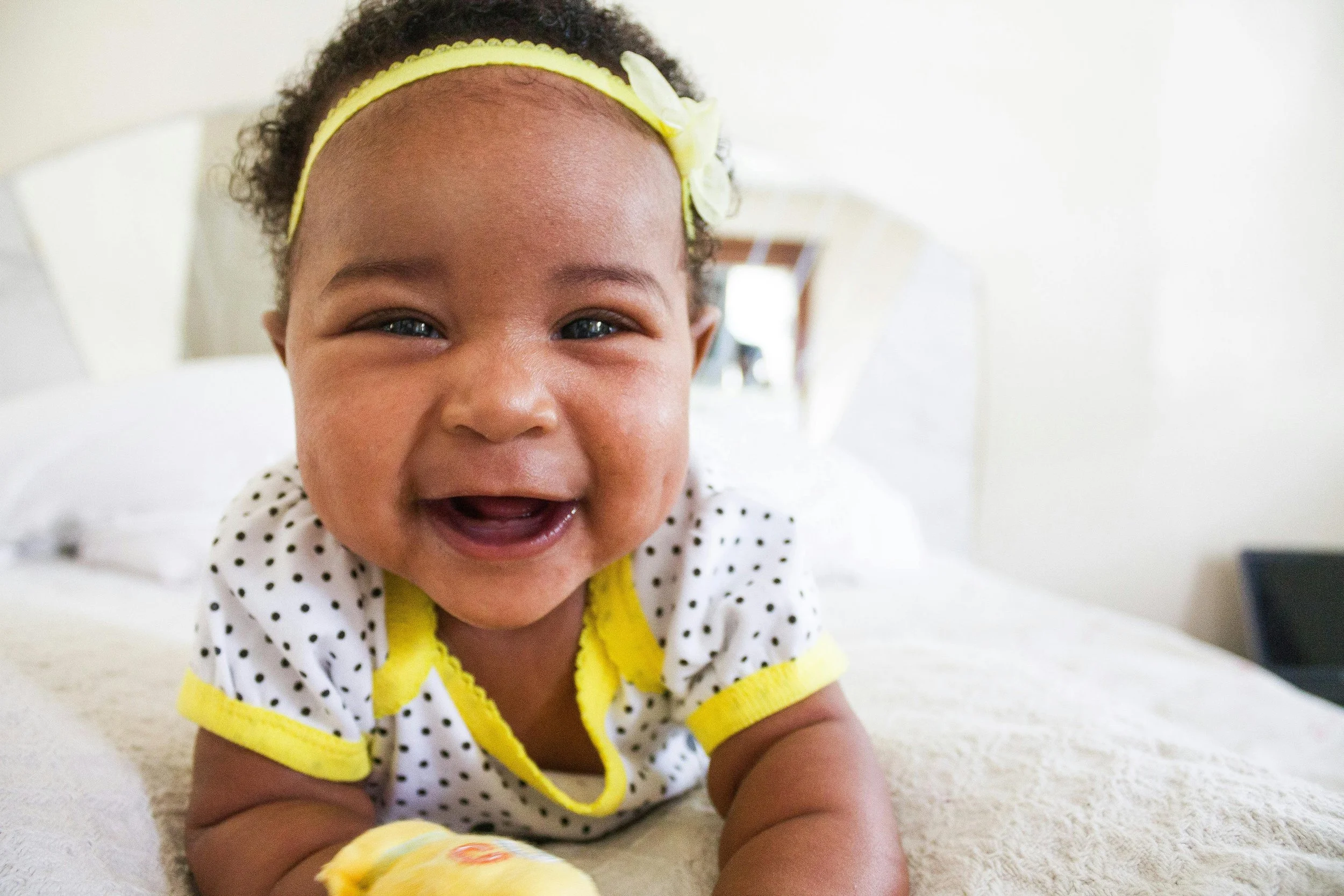 Close-up of a smiling baby girl with blue eyes, wearing a yellow headband with a white bow, lying on her stomach on a bed.