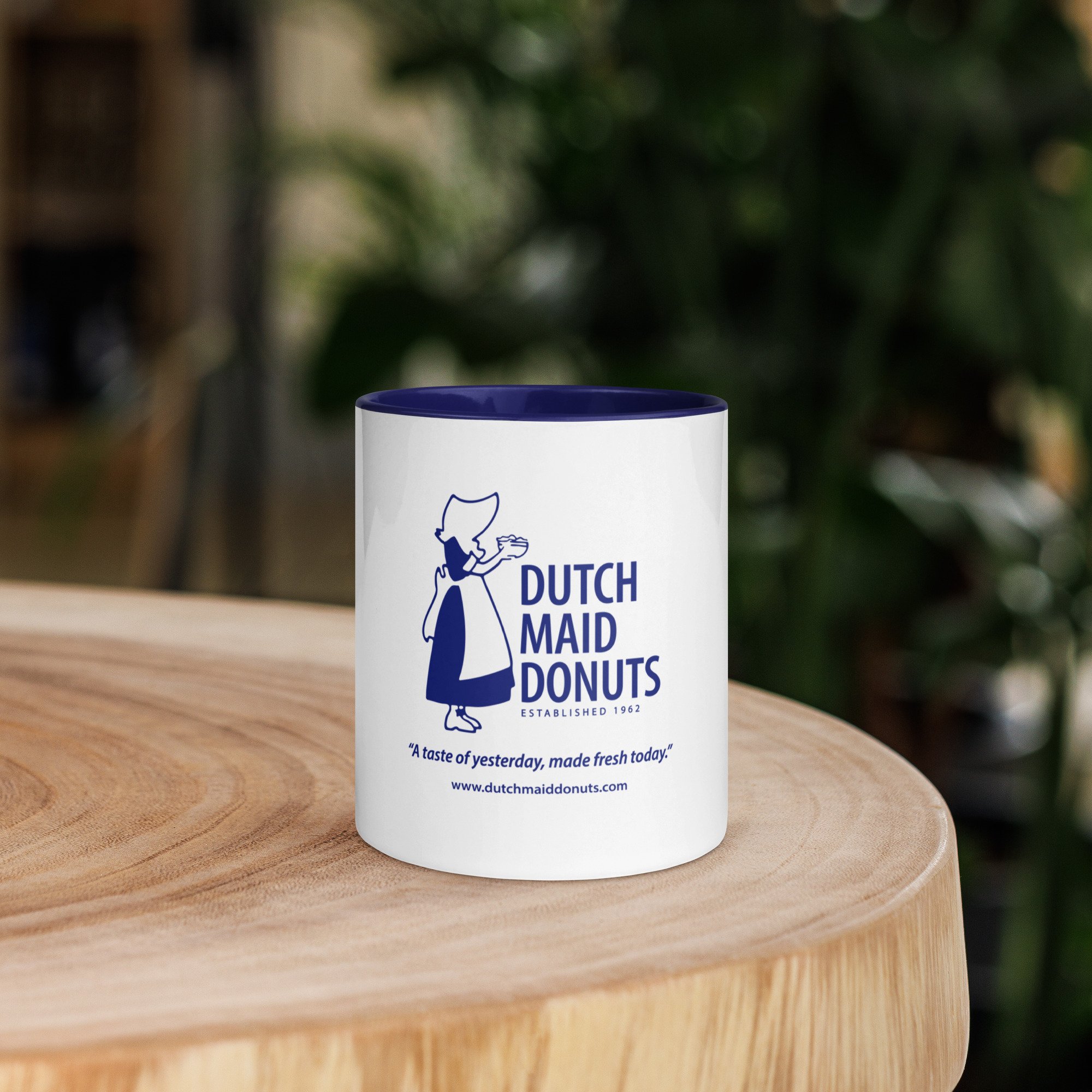 A white mug with blue interior and a logo of a maid holding a tray, reading 'Dutch Maid Donuts', established 1962, with the slogan 'A taste of yesterday, made fresh today.' including website www.dutchmaiddonuts.com, on a wooden surface with a blurred green background.