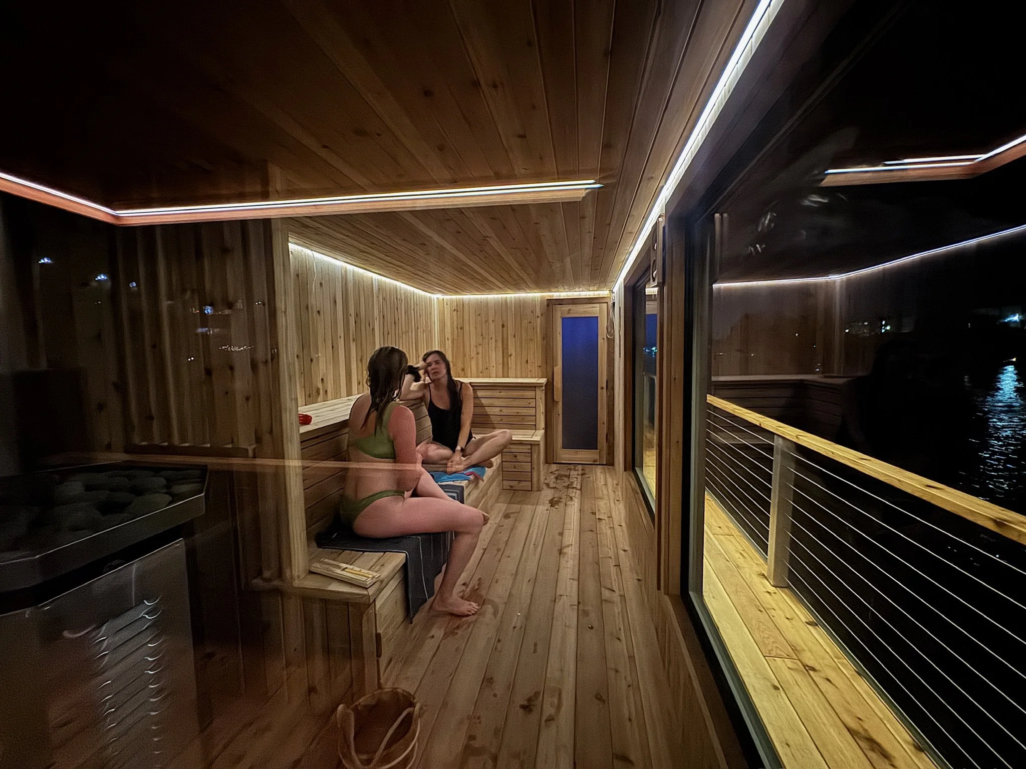 Two women seated on the bench in a lit sauna cabin, the night sky and river visible out the window on the right.