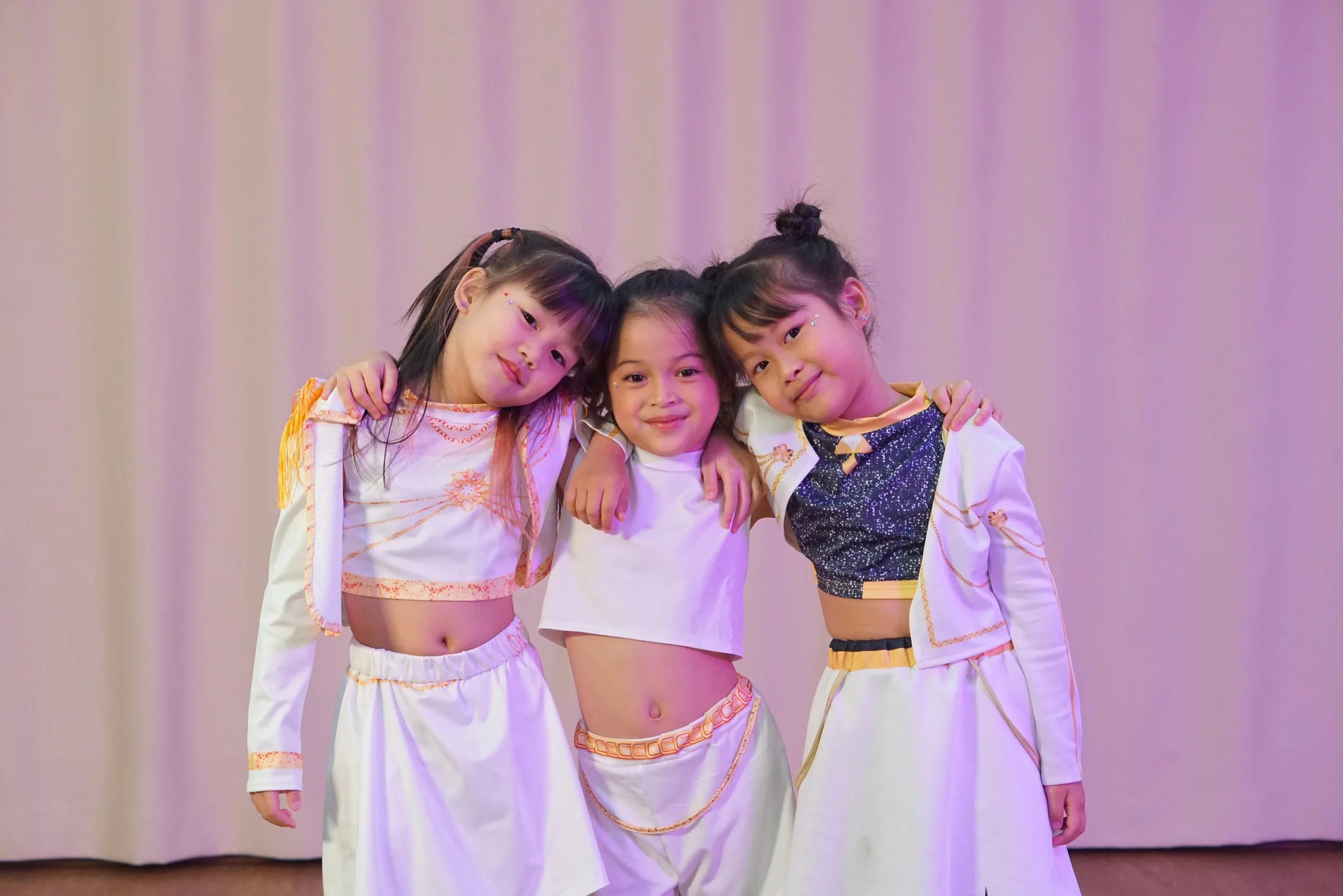 Three young girls in dance costumes hugging and smiling in front of a plain background in Saki Studio Bangkok after a custom course show by Gozen Arts.