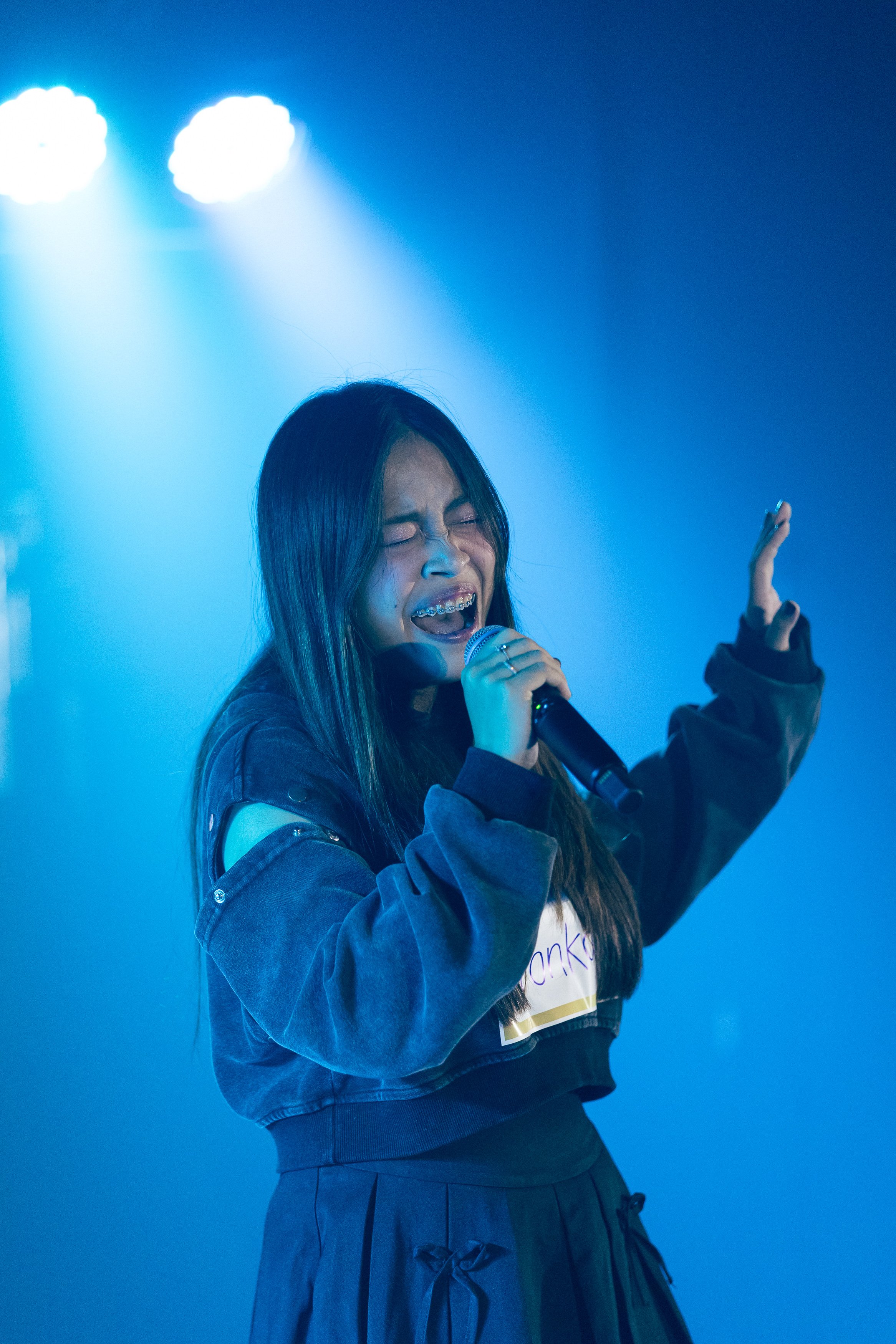 Teacher Hathai of Gozen Arts Bangkok passionately singing into a microphone on stage with blue dramatic lighting.