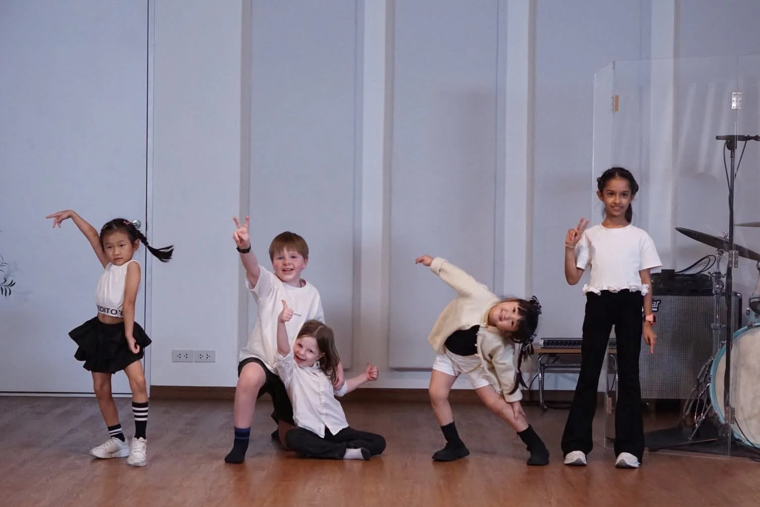 Five young children performing a dance on a wooden floor, with musical instruments and equipment visible in the background. Students of DanSing course of Gozen Arts posing after show in a studio at Bangkok