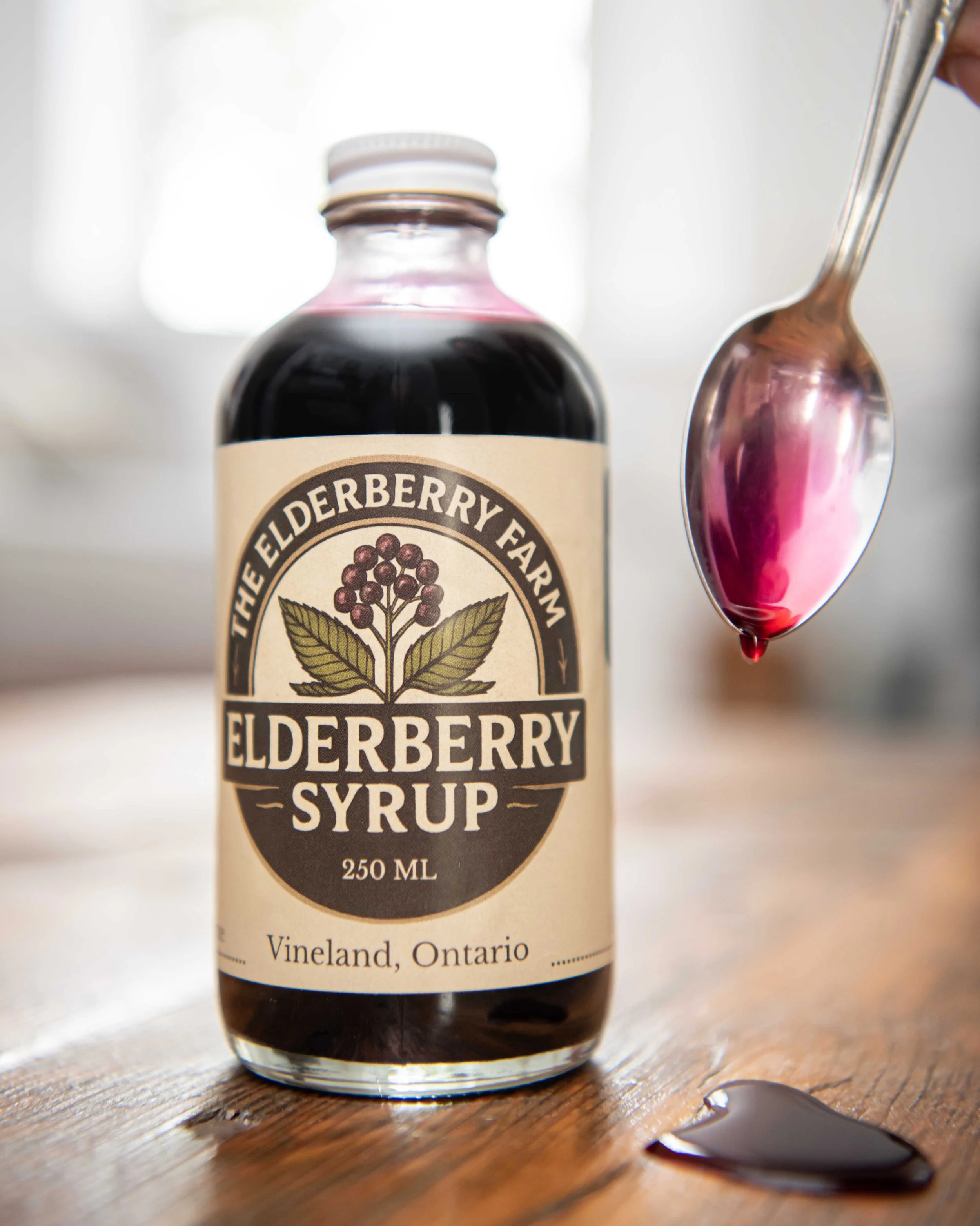 A jar of elderberry syrup next to a spoon with elderberry syrup and a drop of syrup on a wooden surface.