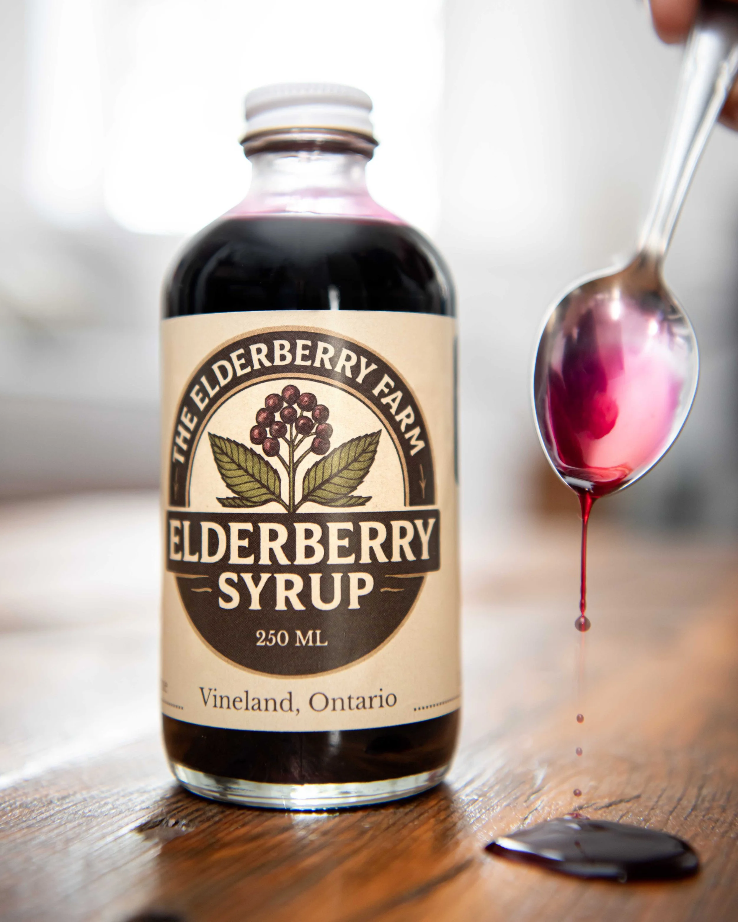 A bottle of elderberry syrup with a spoon showing elderberry syrup dripping off, on a wooden surface.