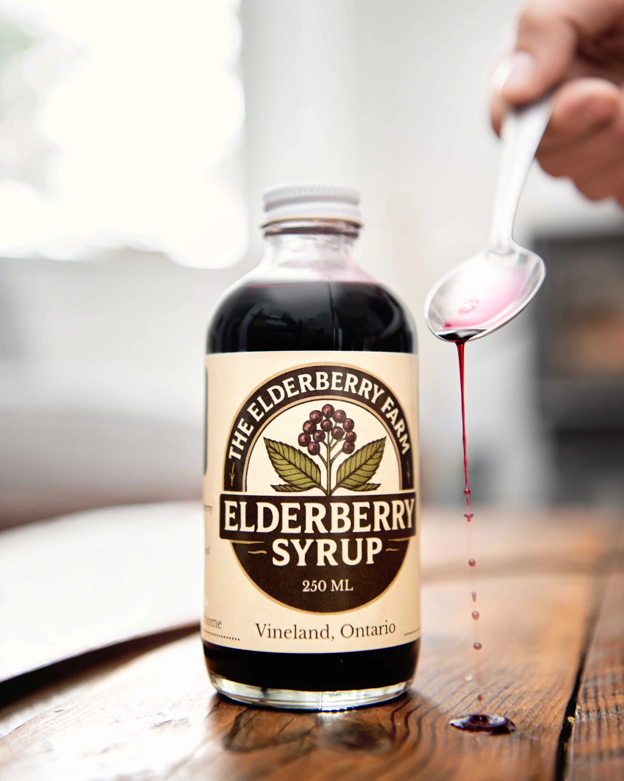 A bottle of elderberry syrup on a wooden surface with a hand holding a spoon dripping elderberry syrup.