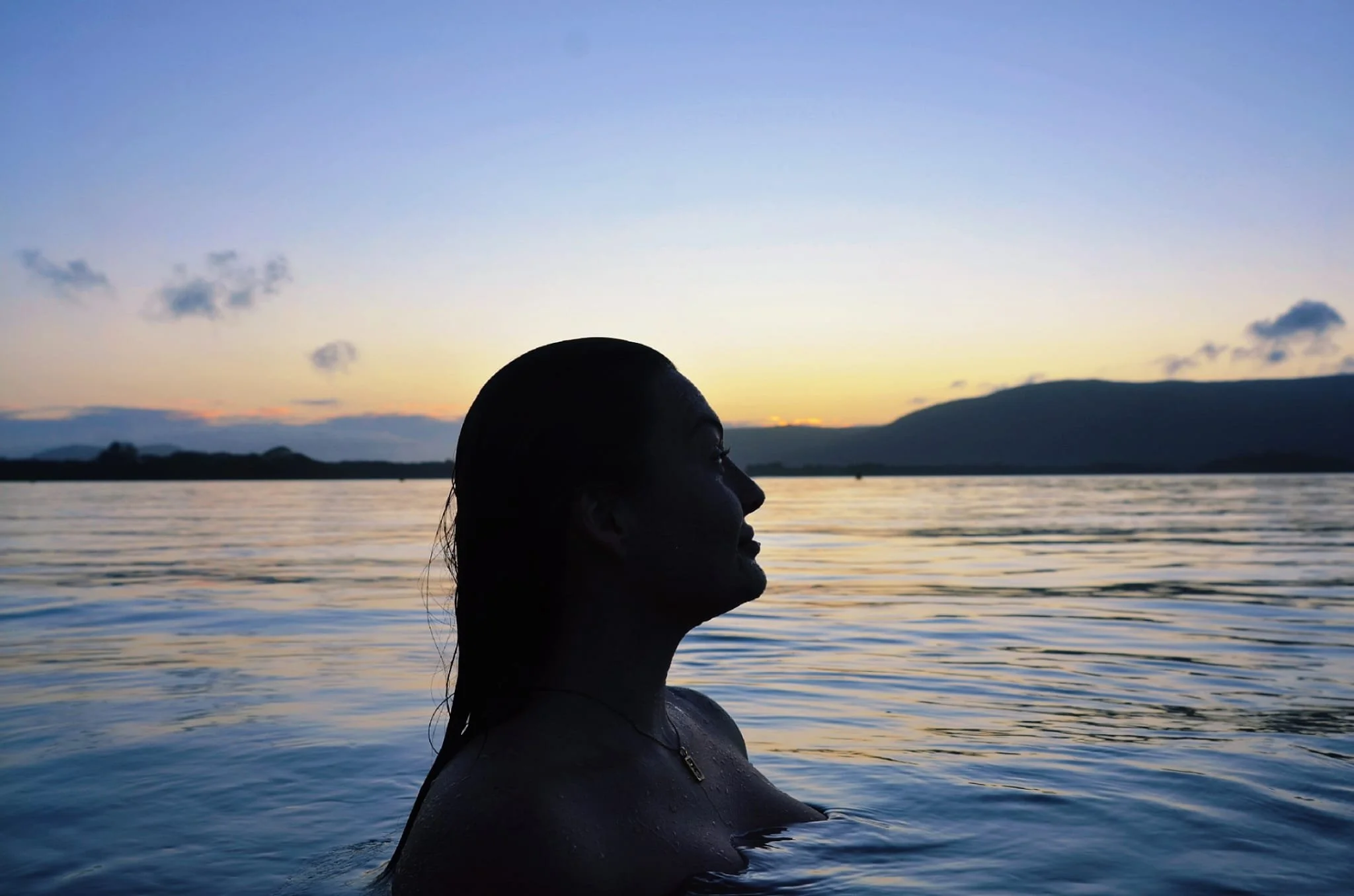 A woman with wet hair is partially submerged in water during sunset, with mountains and a partly cloudy sky in the background.
