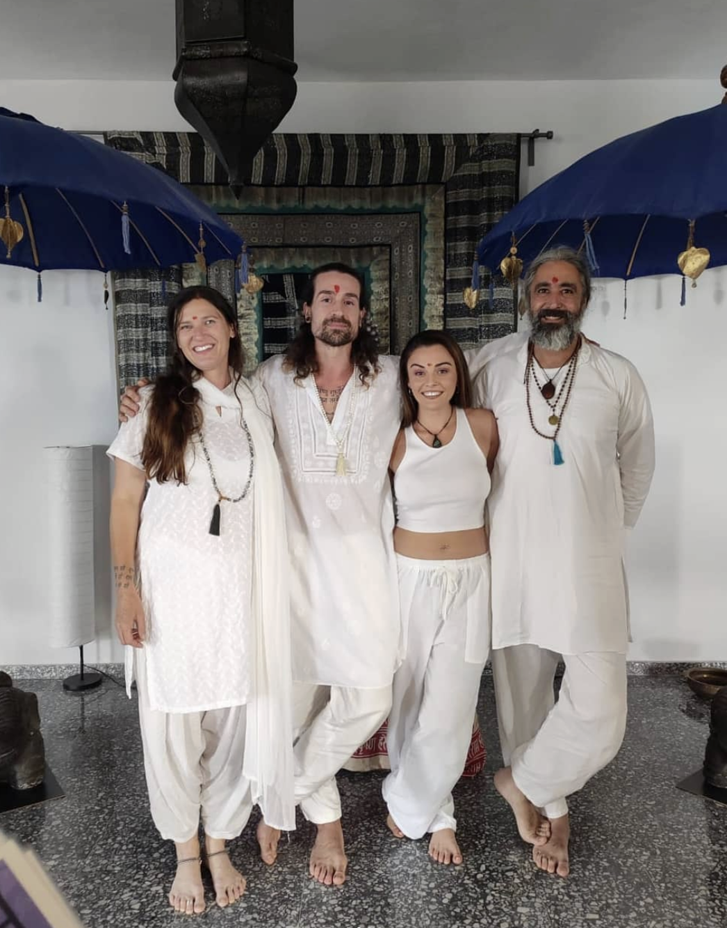 Four people dressed in white traditional Indian clothing, standing together indoors, with two blue umbrellas adorned with hanging gold hearts in the background, and a decorative curtain behind them.