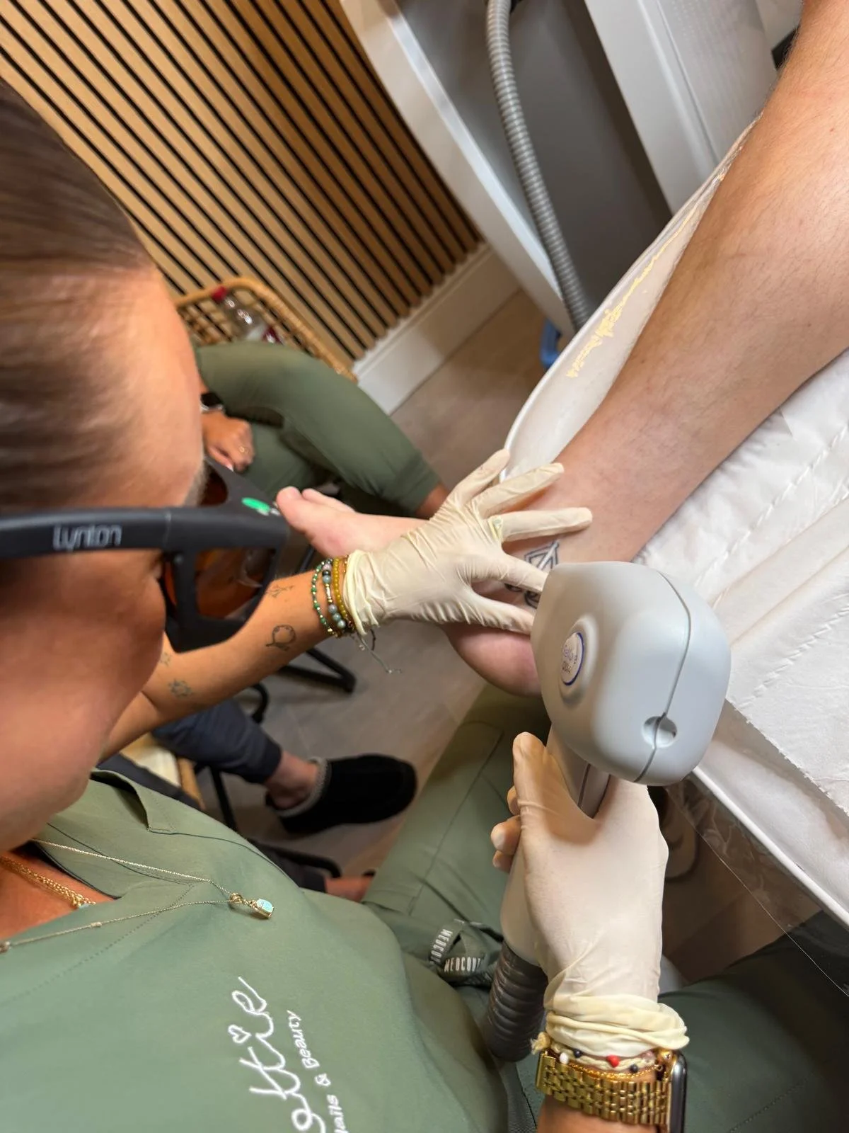 A healthcare worker performs laser hair removal on a person's leg in a clinical setting, wearing gloves and holding a laser device.