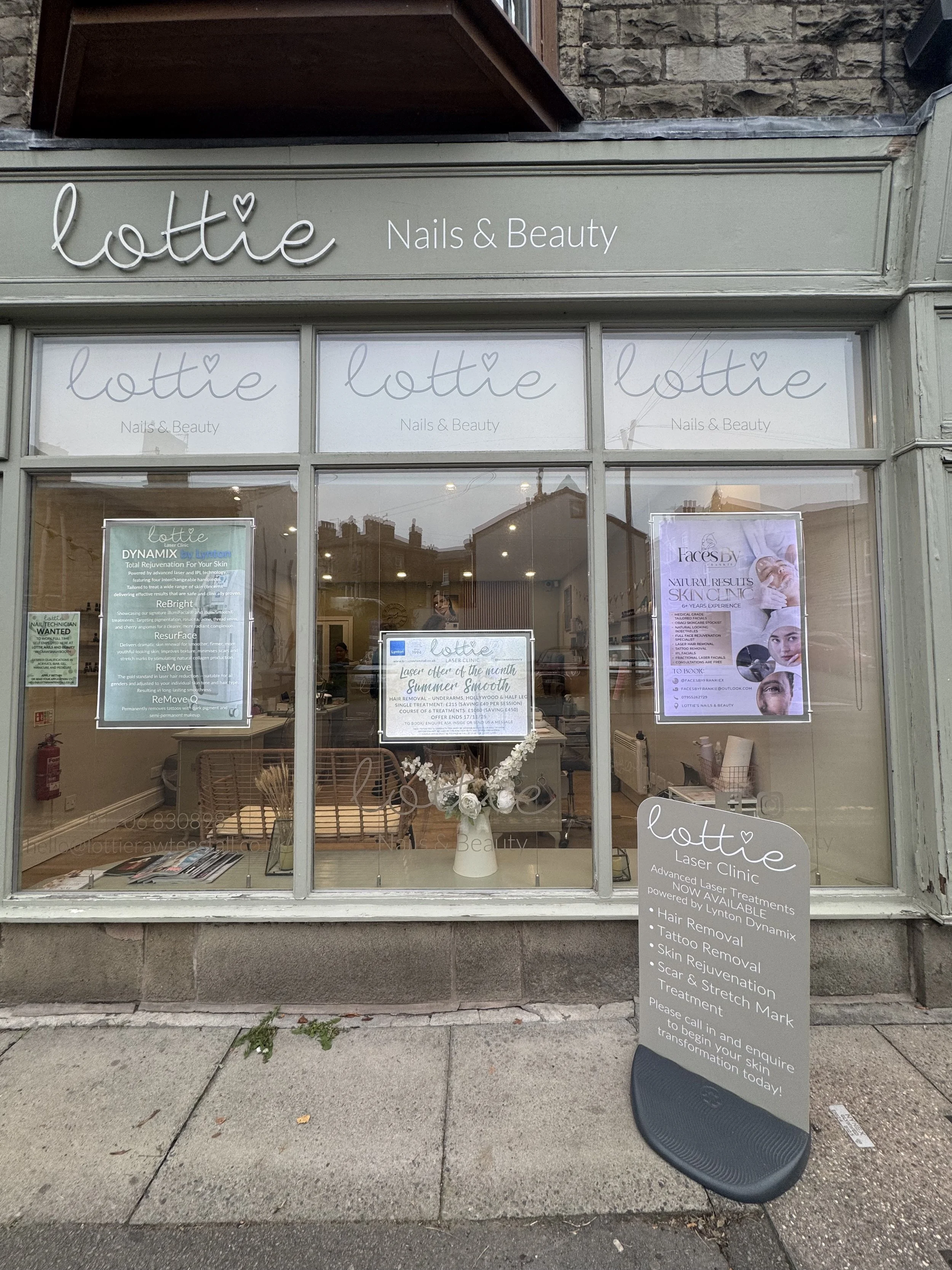 Storefront of Lottie Nails & Beauty salon with large glass windows, signs advertising laser treatments, skincare, and other beauty services, and a sidewalk outside with a signboard and a black chair.