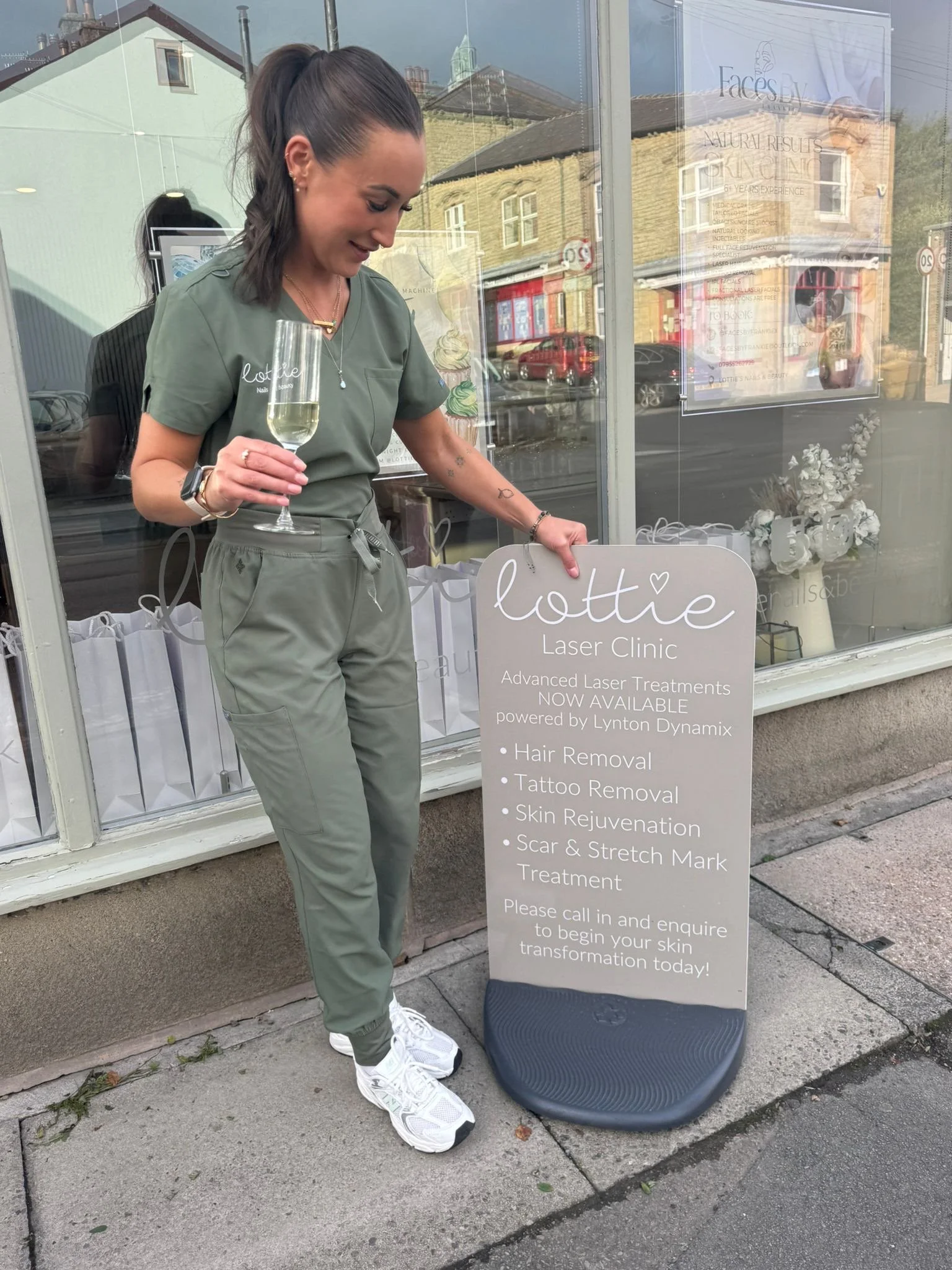 A woman standing outside a laser clinic holding a glass of champagne, wearing green scrubs and white sneakers, with a sign advertising laser treatments like hair removal, tattoo removal, skin rejuvenation, and scar treatment.