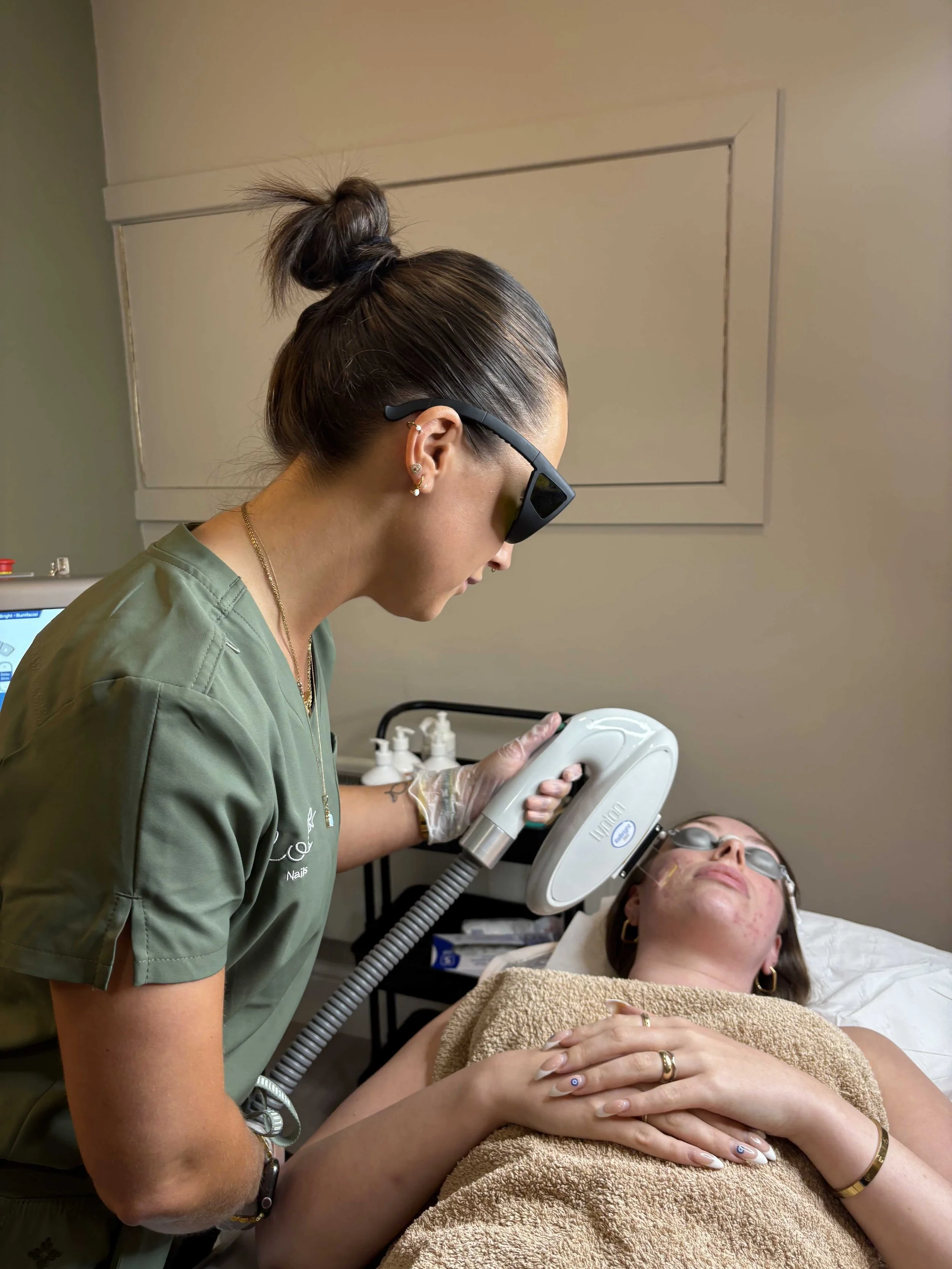 A woman is lying on a medical bed with her hands crossed on her stomach, wearing protective glasses, receiving a laser skin treatment from a technician in a clinic room.