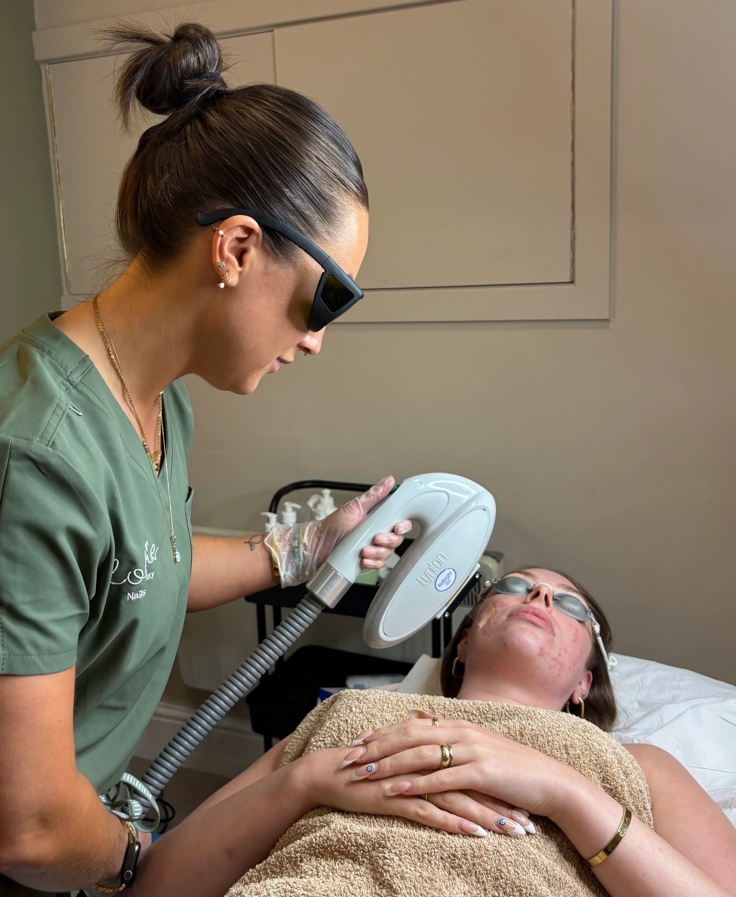 A woman is lying down at a beauty clinic while a technician uses a laser device on her forehead. The woman is wearing protective glasses and has her hands folded on her chest. The technician is wearing green scrubs and gloves, with her hair tied back. The setting appears to be a professional medical or cosmetic treatment room.
