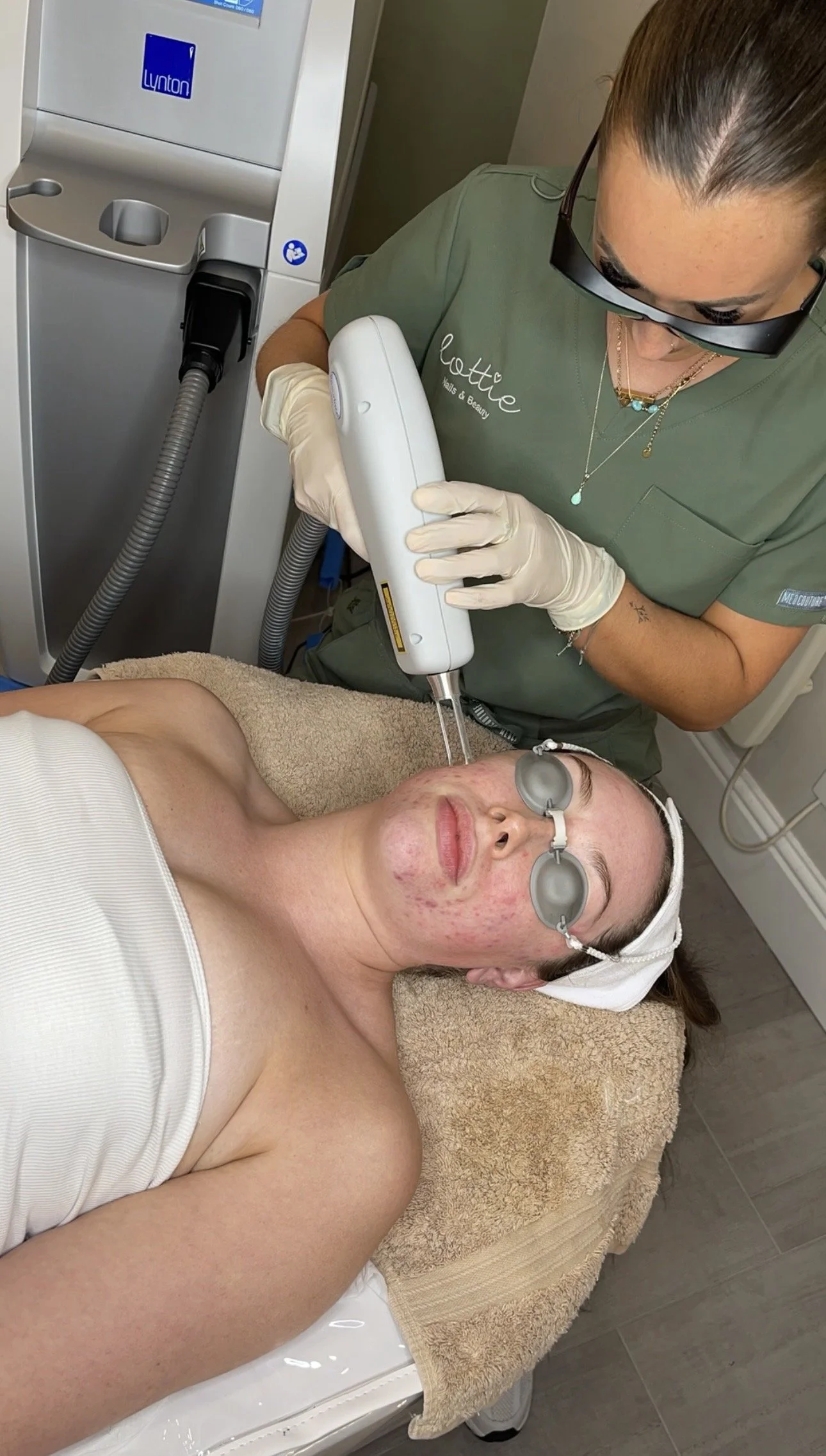 A woman with her eyes covered receiving a cosmetic treatment from a professional in a clinic. The professional is holding a device and wearing gloves. The woman is lying on a treatment bed with a towel underneath her head.