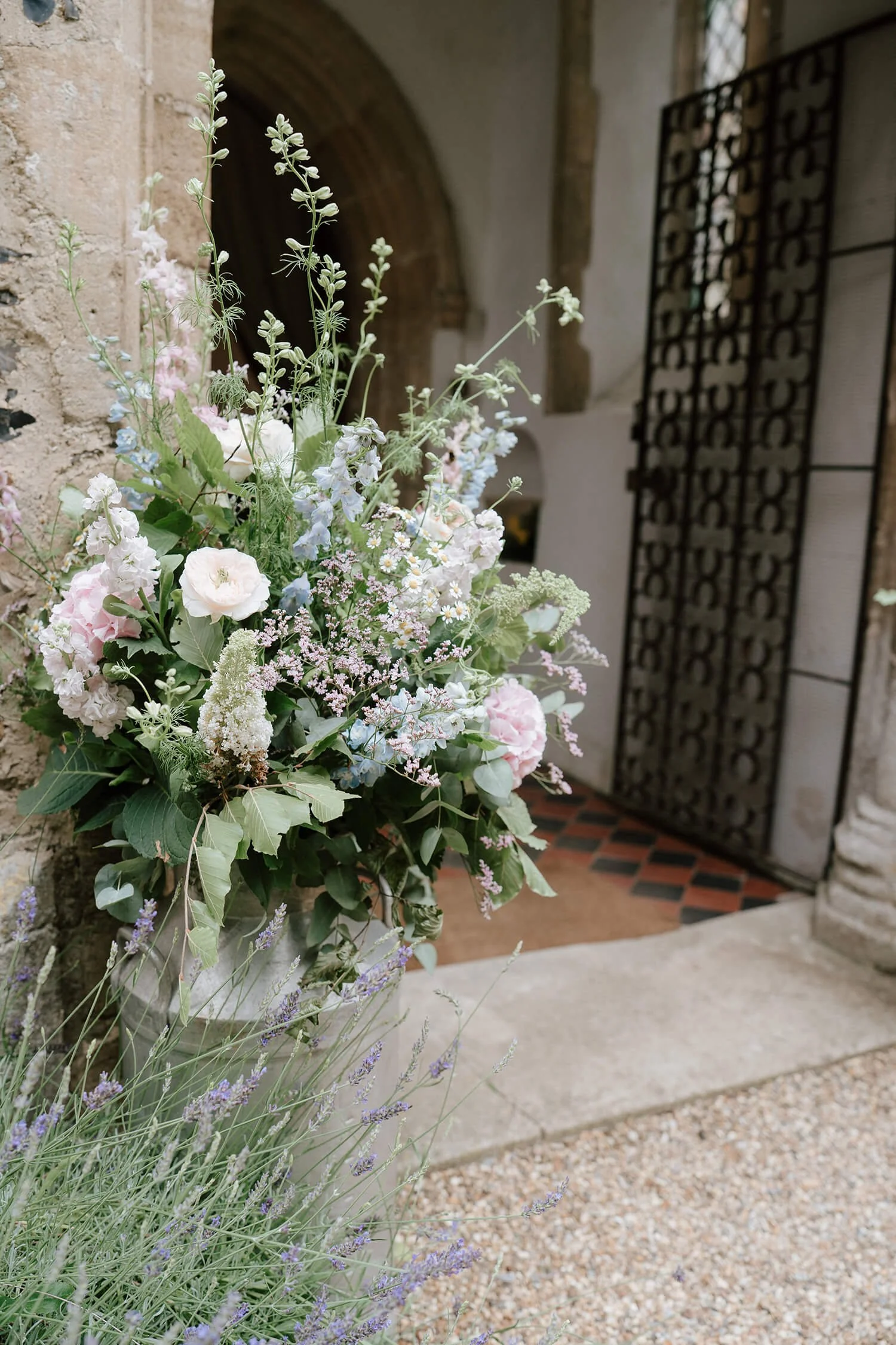 A large flower arrangement with pastel flowers including pink, white, and light blue, placed near a stone wall and entrance with a decorative black iron gate.