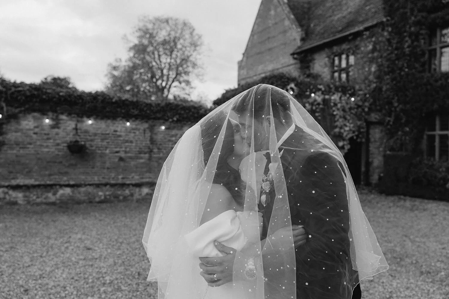 A black and white photo of a bride and groom kissing outdoors, the bride wearing a veil with tiny sparkles, and the groom in a suit, with a house and trees in the background.