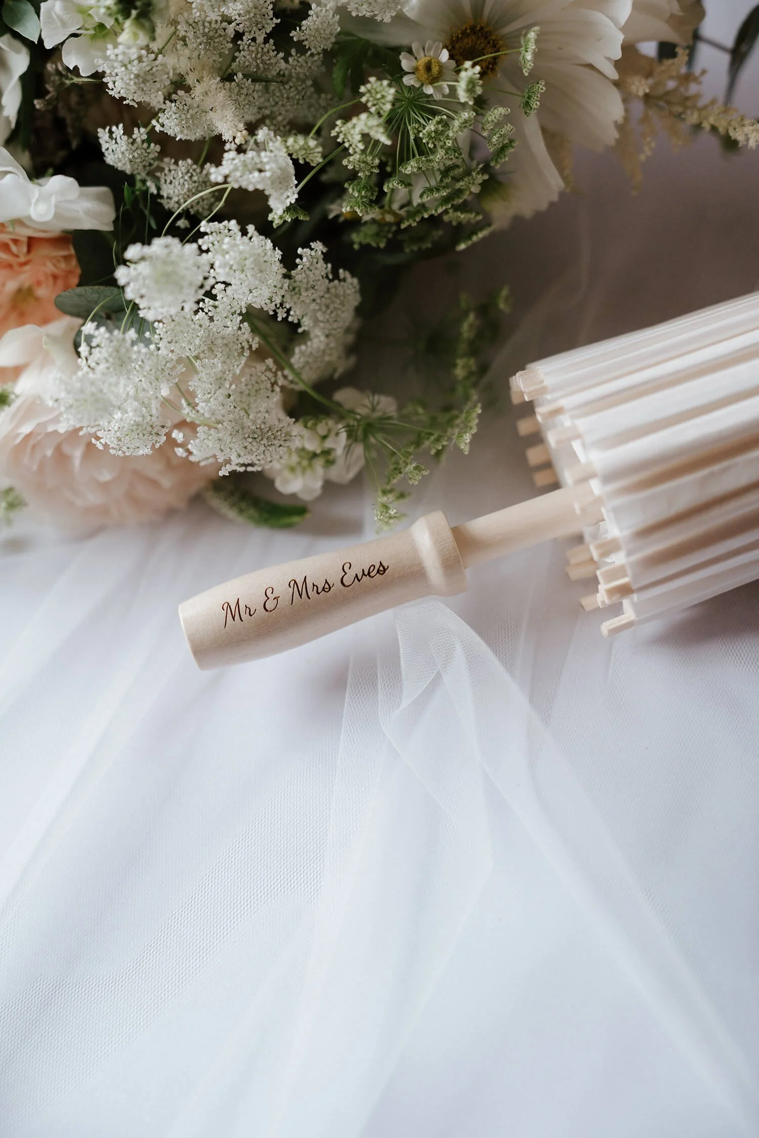 Wedding fan with a wooden handle engraved with 'Mr. & Mrs. Eves' placed on a white fabric with a bouquet of white and pink flowers in the background.