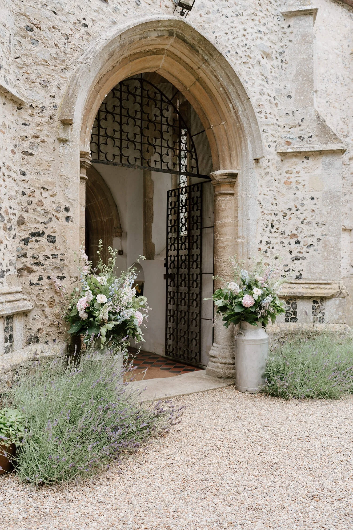 Stone building entrance with arched doorway, decorative iron gate, and flower arrangements in large vases outside.