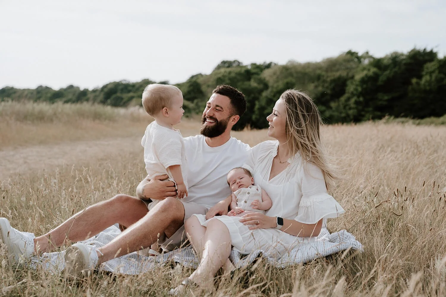 A family of four sitting in a grassy field during daytime, smiling and enjoying each other's company. The father has a beard and is holding a young boy, and the mother is holding a small baby.
