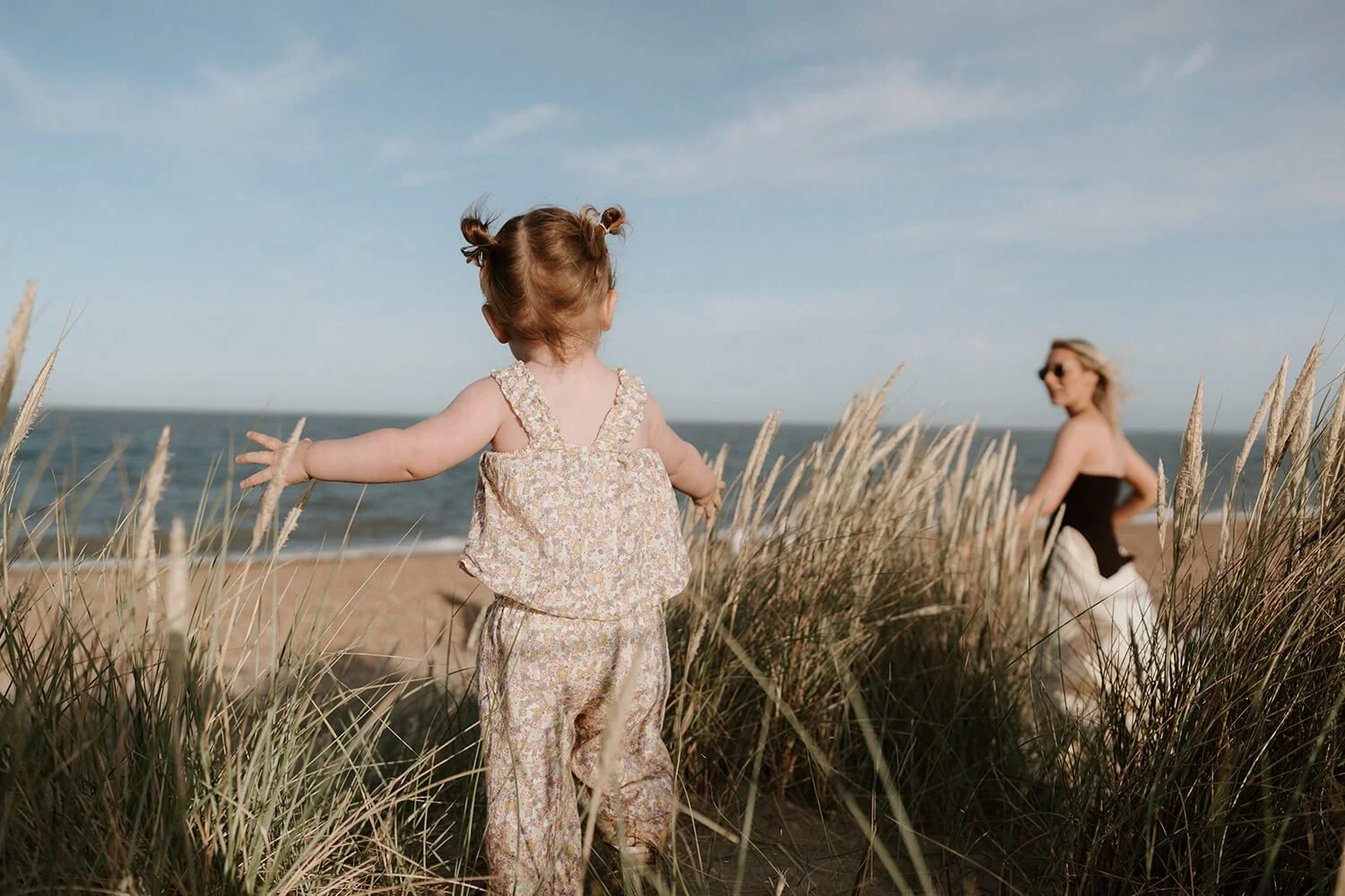 A young girl with curly hair in pigtails standing among beach grass on a sandy beach with her arms outstretched, facing the ocean, with a woman in a black top and white skirt in the background.