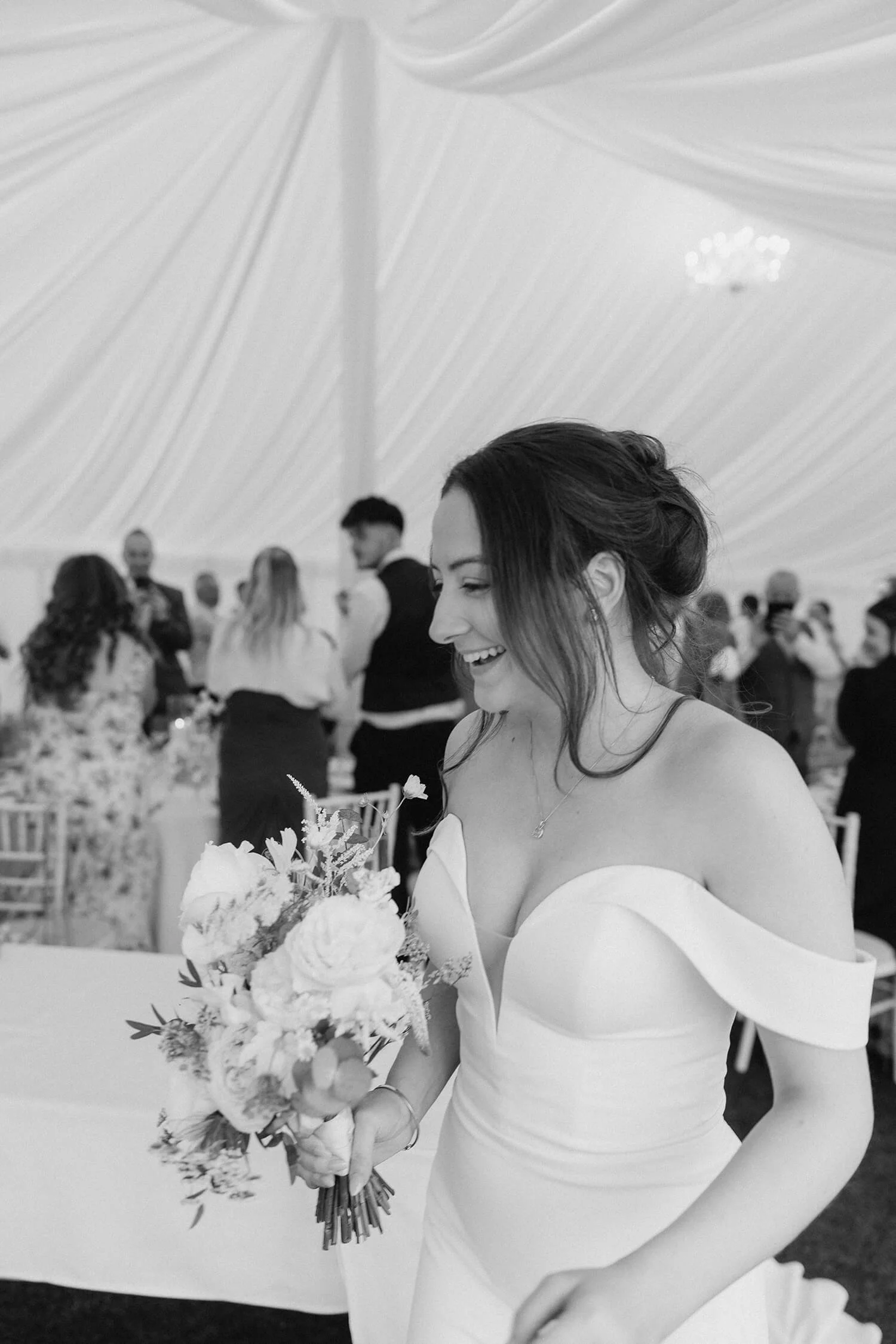 A bride holding a bouquet of flowers at her wedding reception, smiling, inside a large decorated tent.