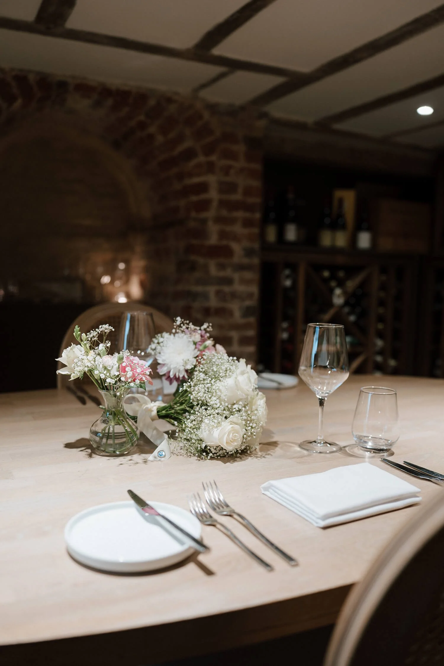 A decorated dining table with a floral centerpiece, wine glasses, a plate with a knife and fork, and neatly folded napkins in a rustic restaurant setting with brick walls and wine bottles in the background.