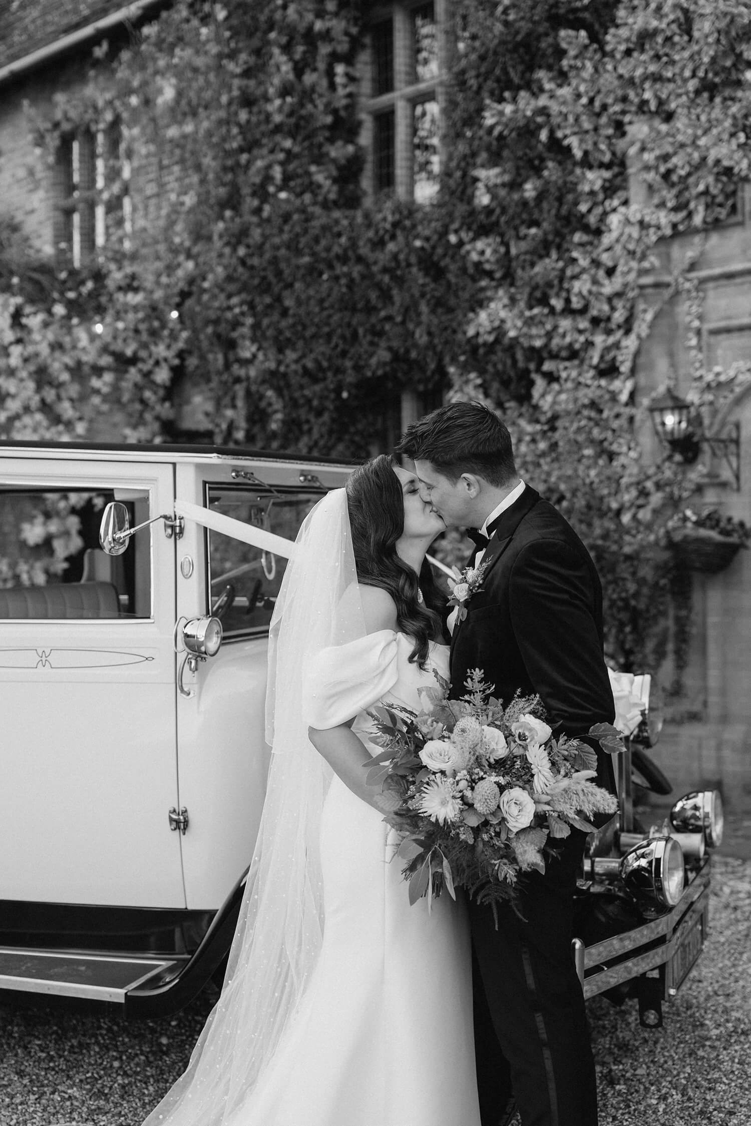 Black and white photo of a bride and groom kissing next to a vintage car, with lush ivy-covered building in the background.