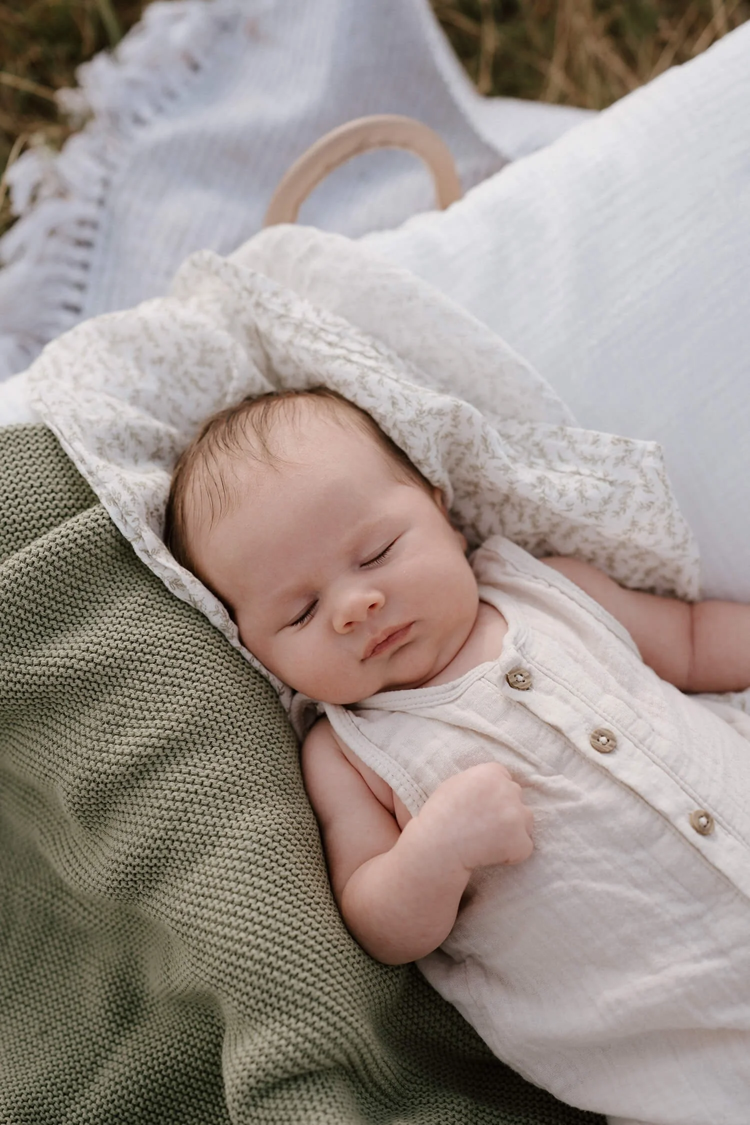A baby sleeping peacefully on a bed with a green knitted pillow and white and beige patterned blankets.