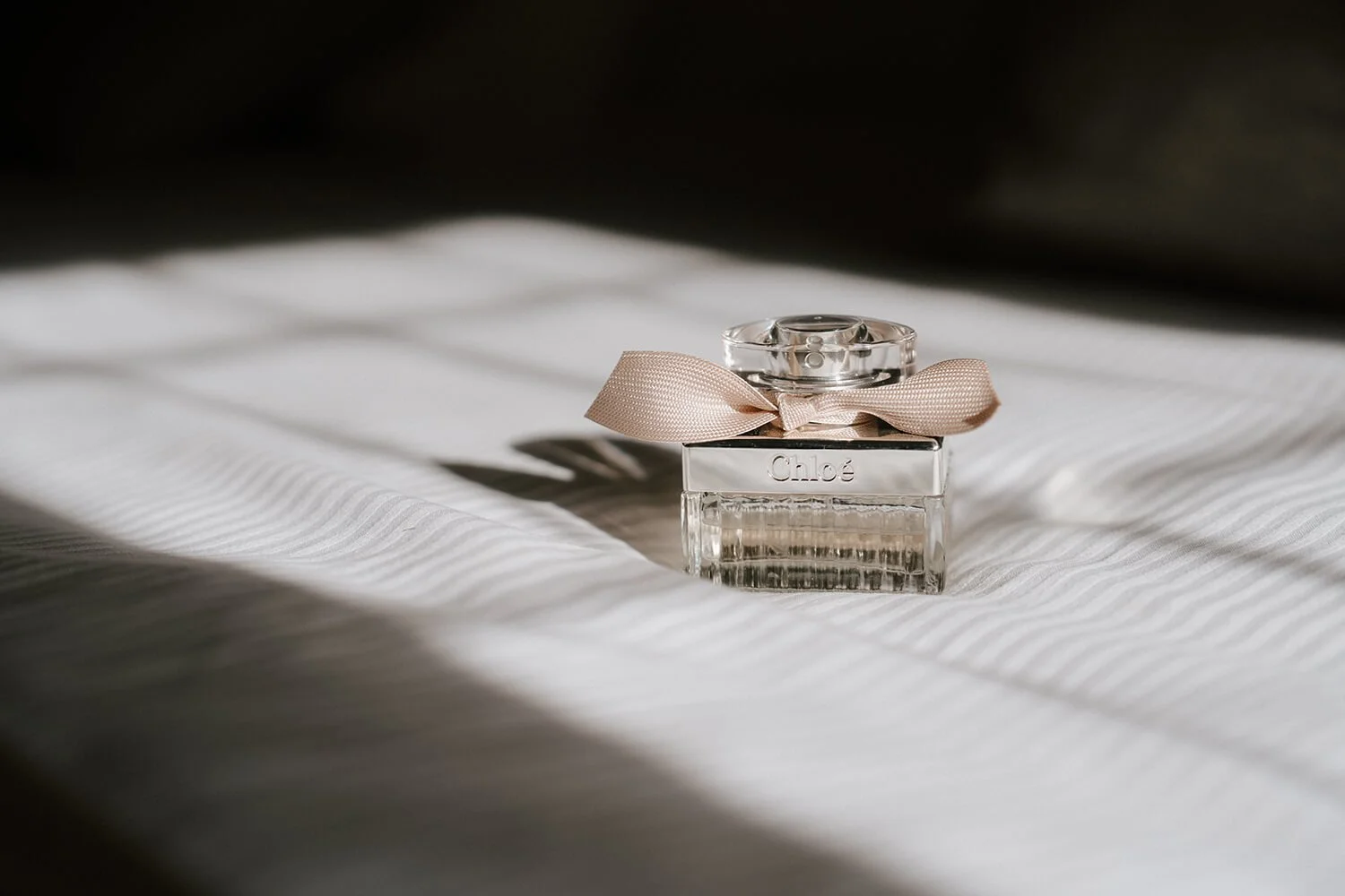 A small, clear glass perfume bottle with a pink ribbon bow on top, sitting on a white, textured fabric surface.
